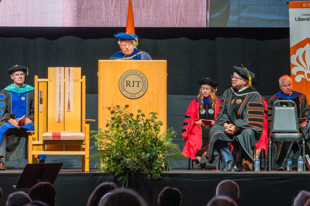 A speaker in academic regalia addresses the audience from the R I T podium while colleagues look on.