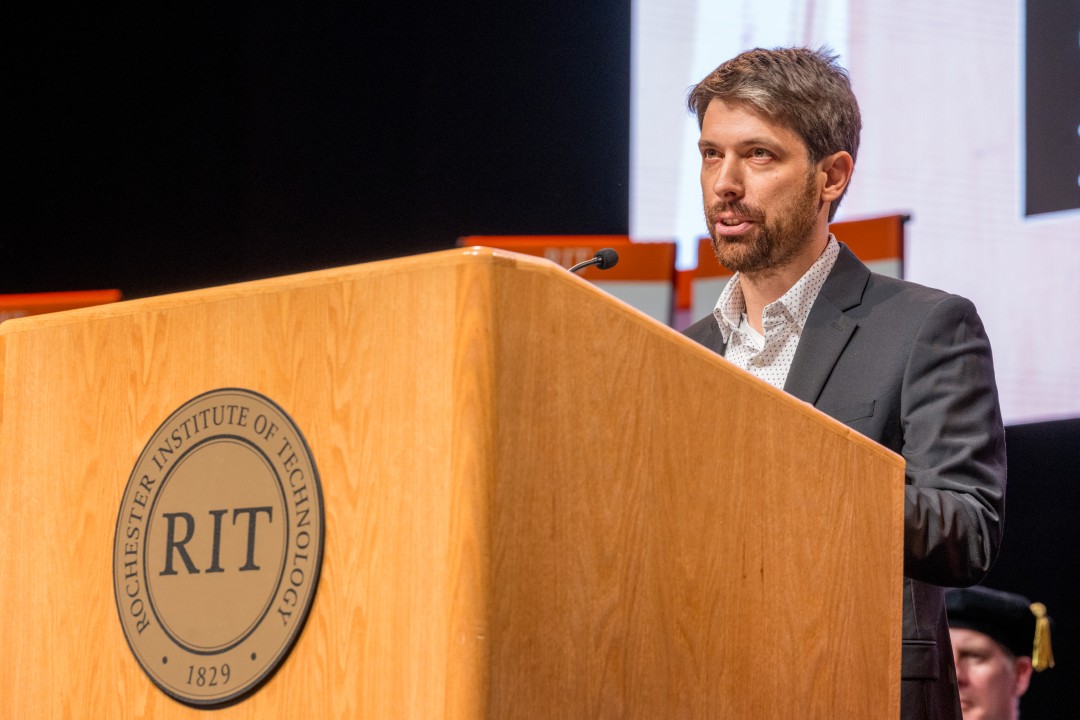 A guest speaker in a suit addresses the audience from the RIT inauguration podium.