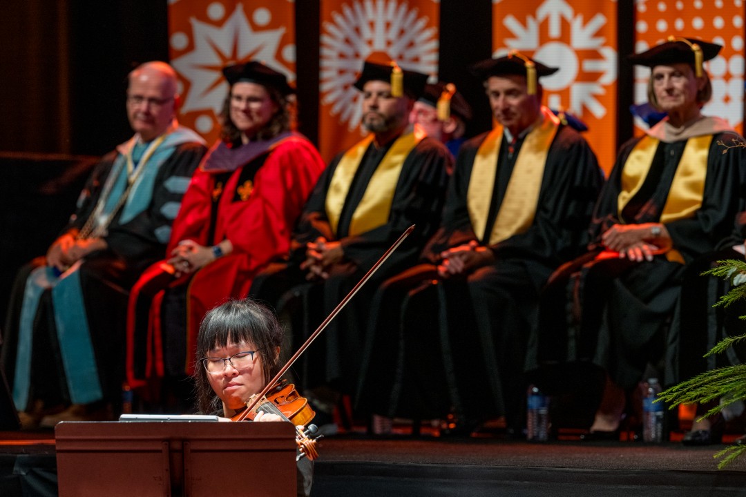 A violinist performs below the stage while faculty in regalia sit in the background.