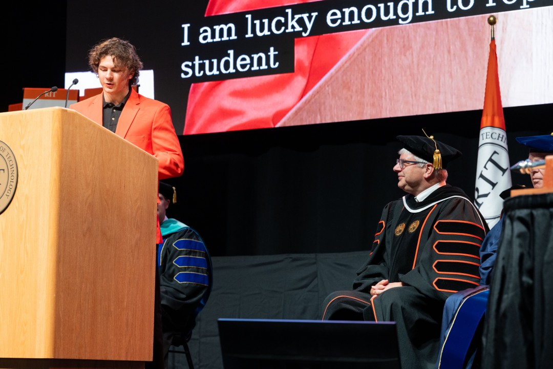 A student in an orange jacket speaks at the podium while the president in regalia listens seated on stage.