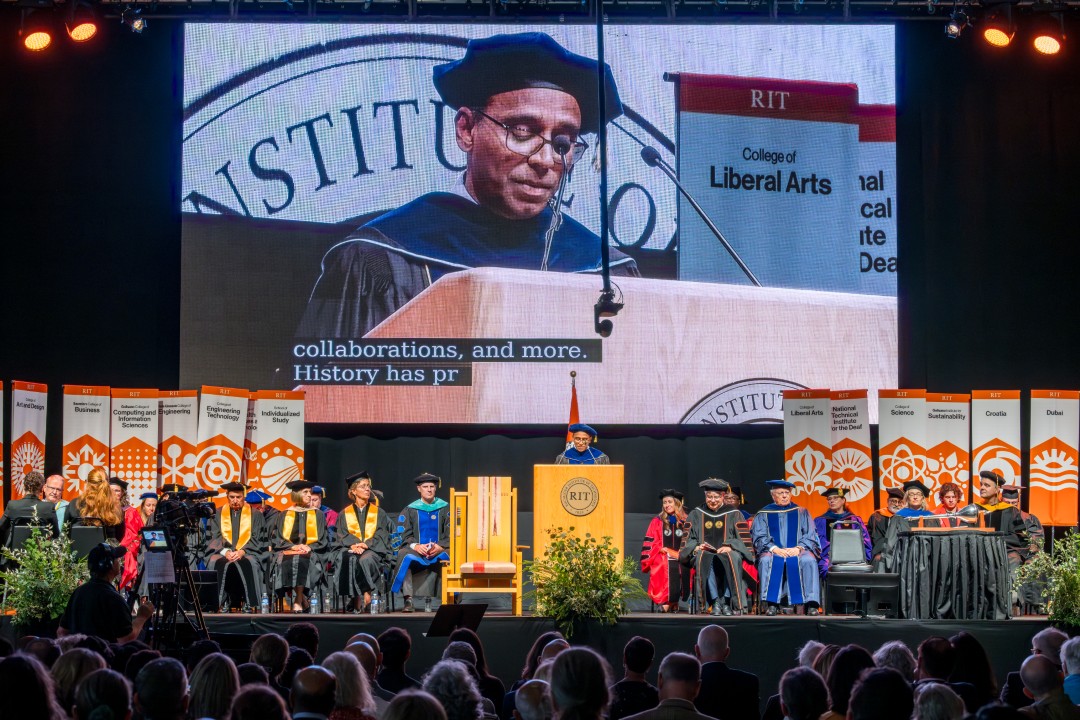 A wide view of the R I T provost delivering remarks at the podium while faculty and trustees sit on stage.