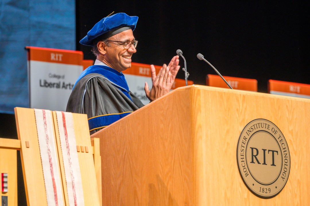 A speaker in academic regalia applauds at a podium bearing the Rochester Institute of Technology seal.