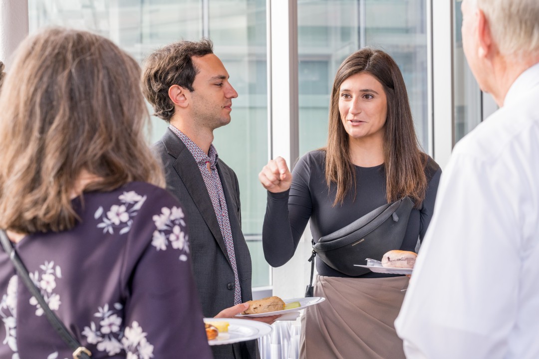Four people stand chatting at a recption event while holding plates of food.
