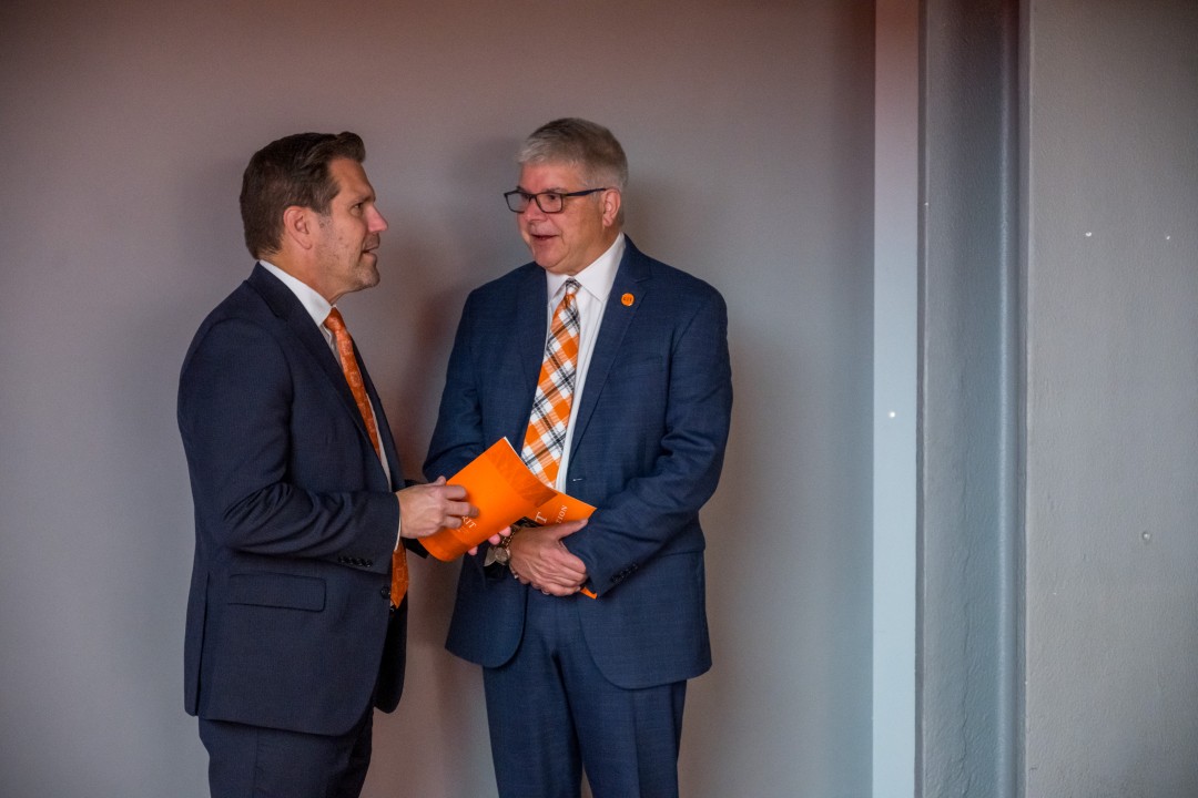 Two men in suits talk while holding orange folders in a hallway at an event.