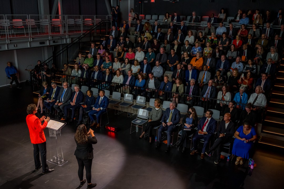 A speaker in an orange suit jacket addresses a large seated audience in an auditorium.