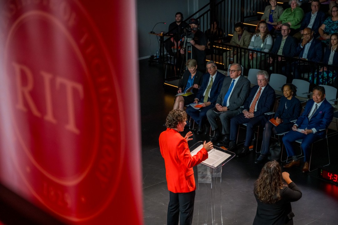 A speaker in an orange suit jacket presents on stage in front of seated attendees with an R I T banner in the foreground.