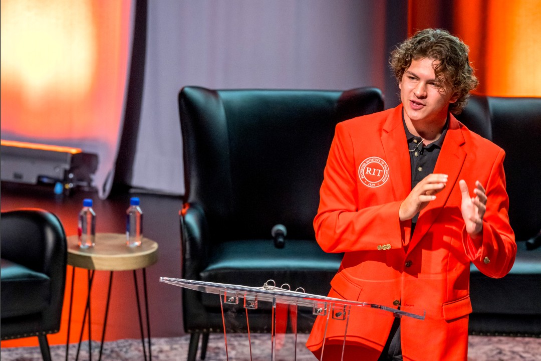 The R I T student government president wearing an orange suit jacket speaks at a clear podium on stage.