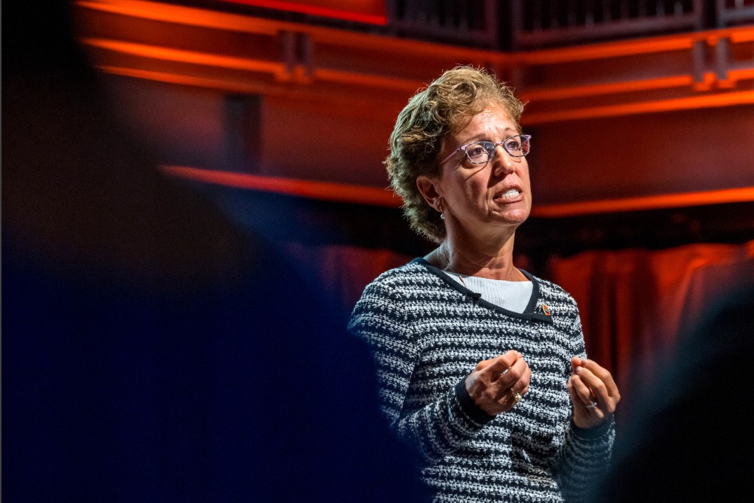 A woman wearing glasses and a striped sweater speaks on stage.