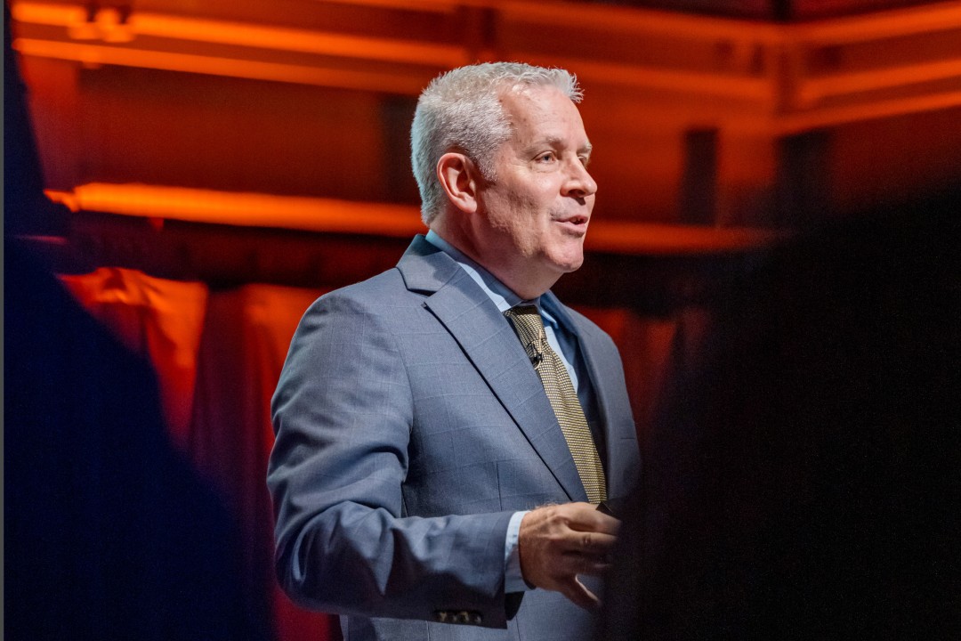 A man in a gray suit and tie gives a talk on stage under warm lighting.