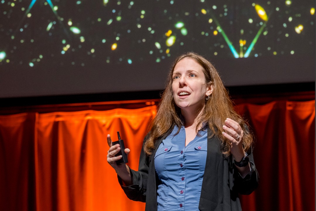 A woman in a blue blouse and black jacket speaks in front of a projection of galaxies.
