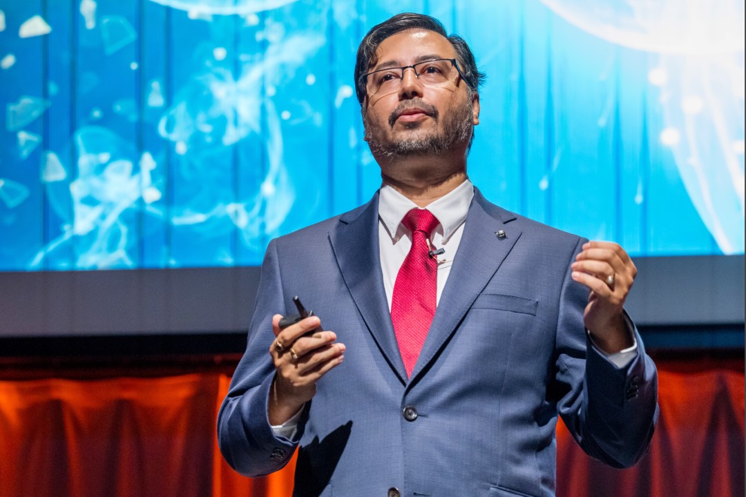 A man in a blue suit and red tie gestures while speaking on stage in front of a blue science-themed background.