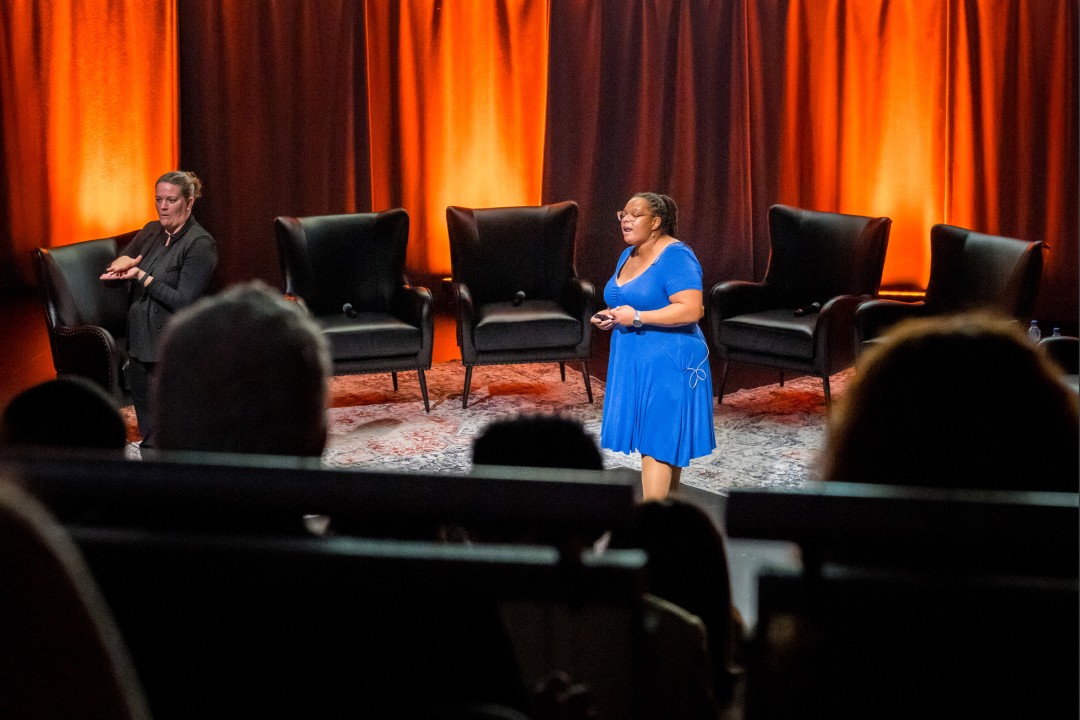 A wide view of a stage surrounded by armchairs, with a woman in the center giving a presentation, and a sign langage interpreter off to the side.