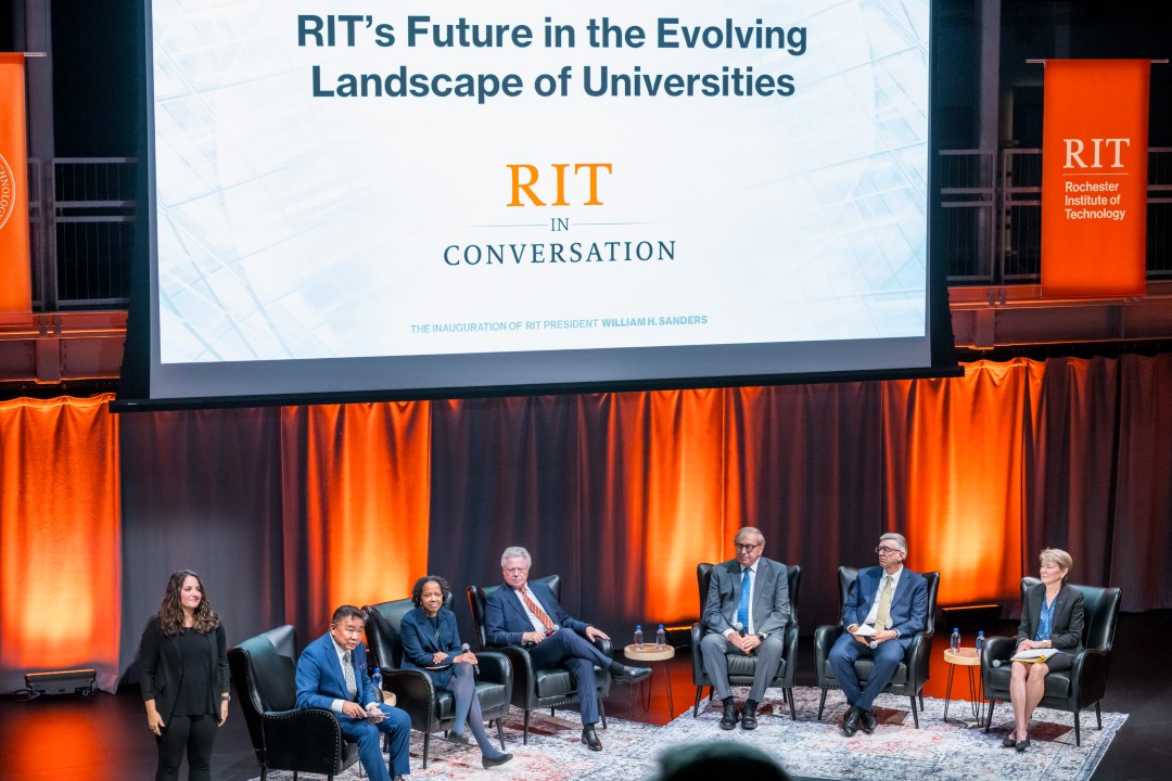 A stage with six panelists seated, a sign language interpreter off to the side, and a large projector screen showing a slide that reads 'RIT's Future in the Eveolving Landscape of Universities - R I T in conversation.'