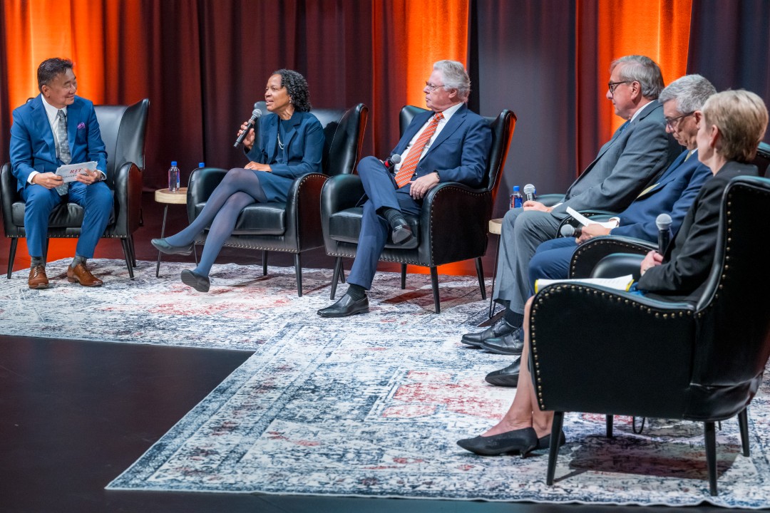 One of the six panelists speaking into a microphone as the others listen.