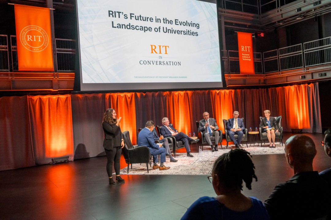 A view from behind the first row of audience members of a stage with six panels seated, a sign language interpreter off to the side, and a large projector screen showing a slide that reads 'RIT's Future in the Eveolving Landscape of Universities - R I T in conversation.'