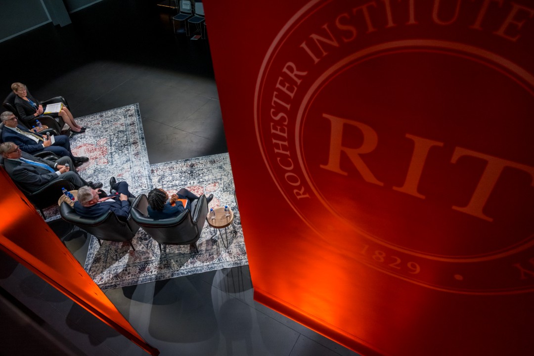 An overhead view of the speakers on the stage, with an orange R I T banner uplit in the foreground.