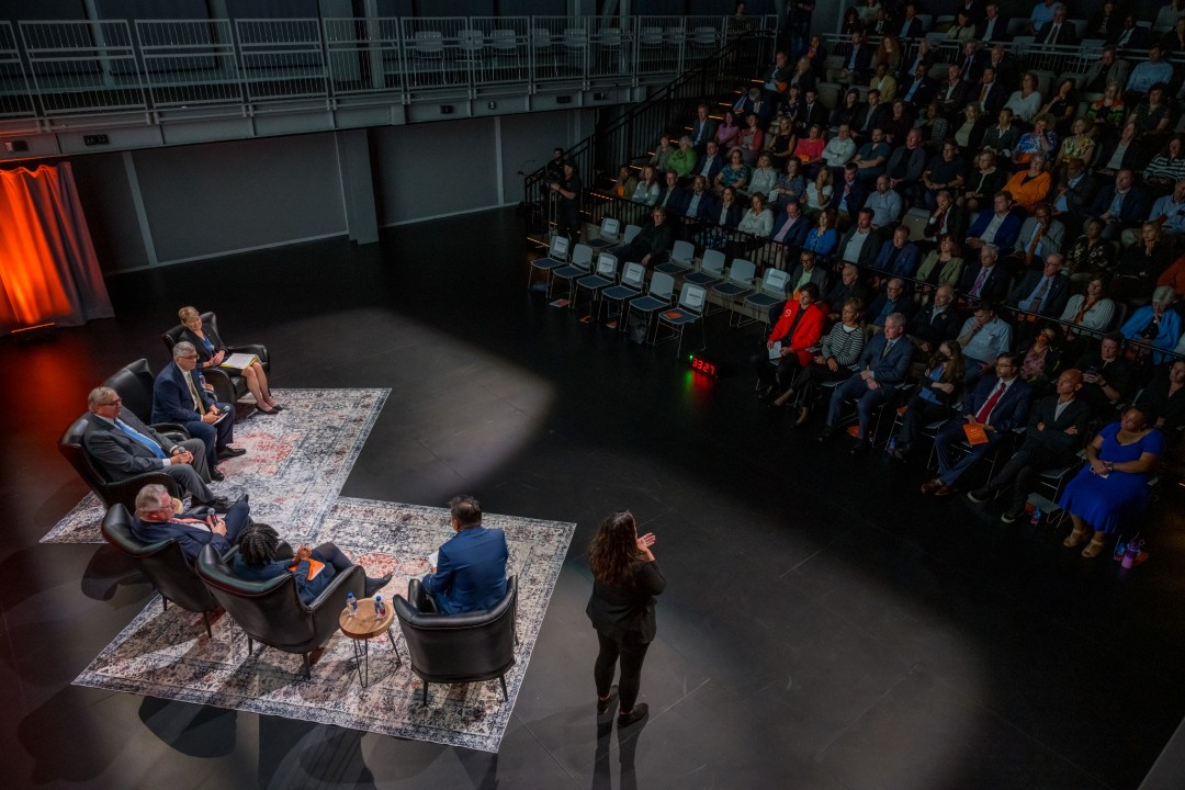 An overhead view of the six panelists and several rows of audience members seated in an auditorium.