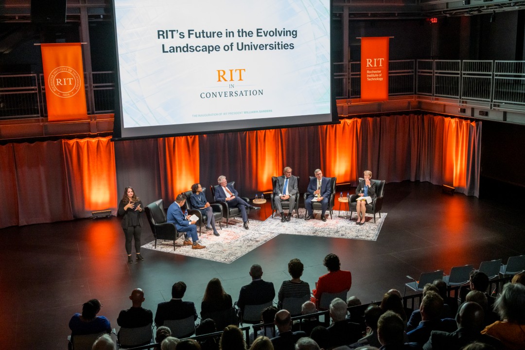 A stage with six panelists seated, a sign language interpreter off to the side, and a large projector screen showing a slide that reads 'RIT's Future in the Eveolving Landscape of Universities - R I T in conversation.'