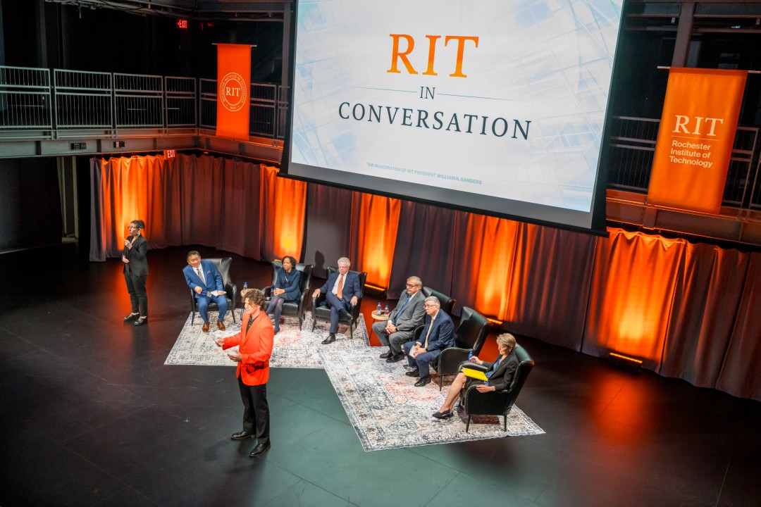 A view looking down onto the stage of six panelists, with one standing speaker in the center and a sign language interpreter off to the side.