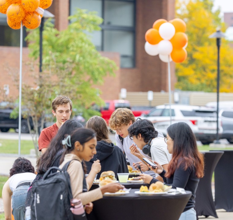 Students gather around a tall table outdoors eating food under balloons in R I T colors.