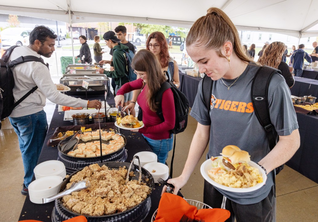 Students serve themselves food from buffet trays while holding plates at a picnic.