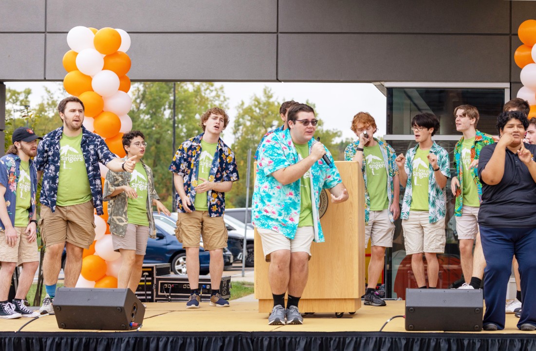 A student singing group in green T-shirts and Hawaiian shirts performs on an outdoor stage with balloons.