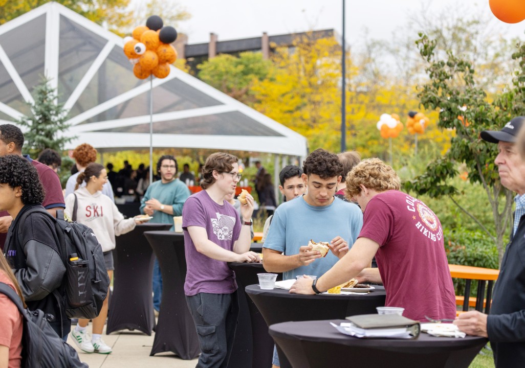 Students eat and socialize at tall tables outside near a tent decorated with balloons.