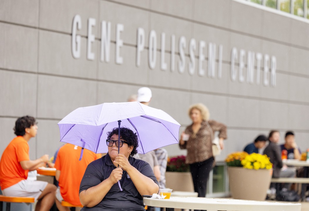 A person sits at a table holding a lavender umbrella in the rain outside the Gene Polisseni Center.