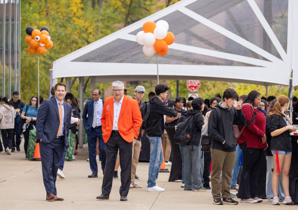 R I T president Sanders walks near a crowd gathered under a tent decorated with orange and white balloons.