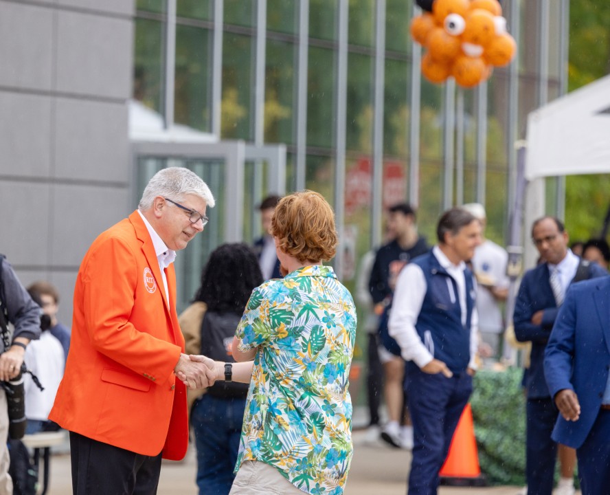 R I T president Sanders in an orange blazer shakes hands with a student in a floral shirt outside an event tent.
