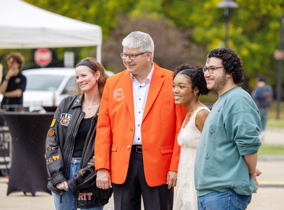 R I T president Sanders in an orange blazer poses for a group photo with three students outdoors.