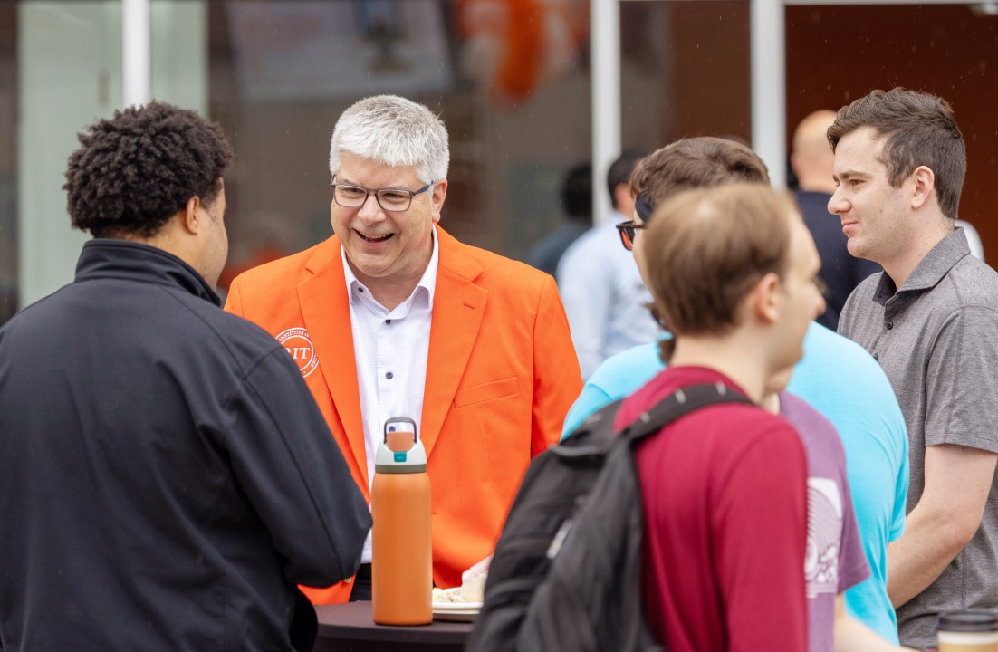 R I T president Sanders in an orange blazer laughs while talking with students around a tall table.
