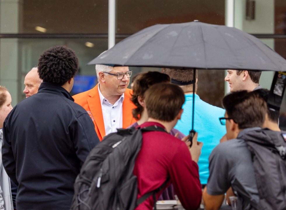 A group of students with backpacks gather around R I T president Sanders while one student holds an umbrella.