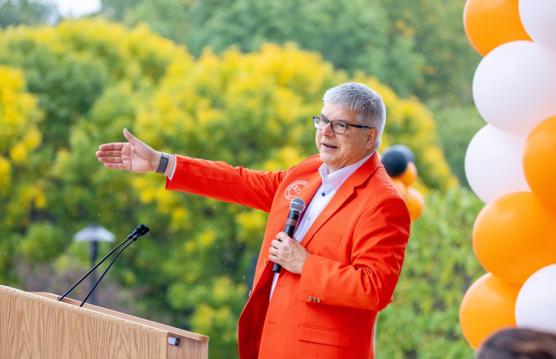 R I T president Sanders in an orange blazer speaks at a podium with a microphone, gesturing beside orange and white balloons.