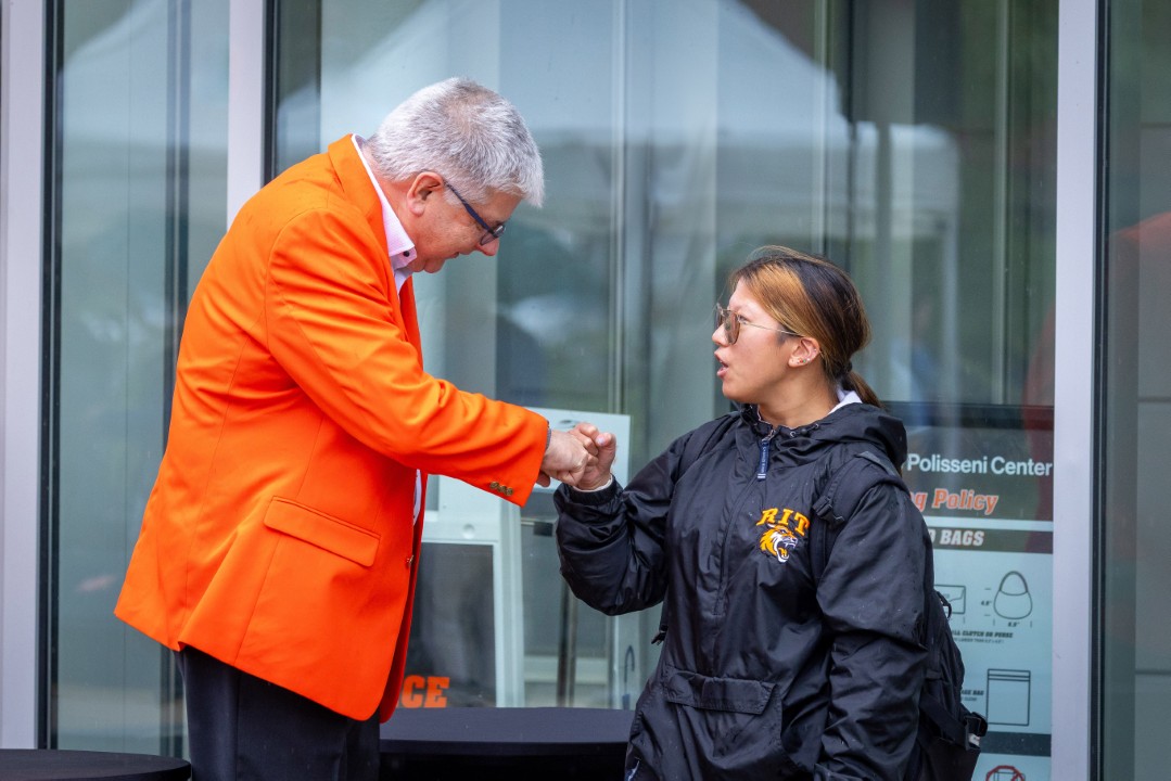 R I T president Sanders in an orange blazer fist bumps a student in a black R I T jacket outside the Polisseni Center.