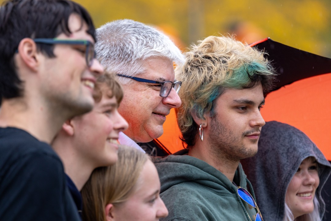 R I T president Sanders in an orange blazer smiles while posing for a close-up photo with a group of students.