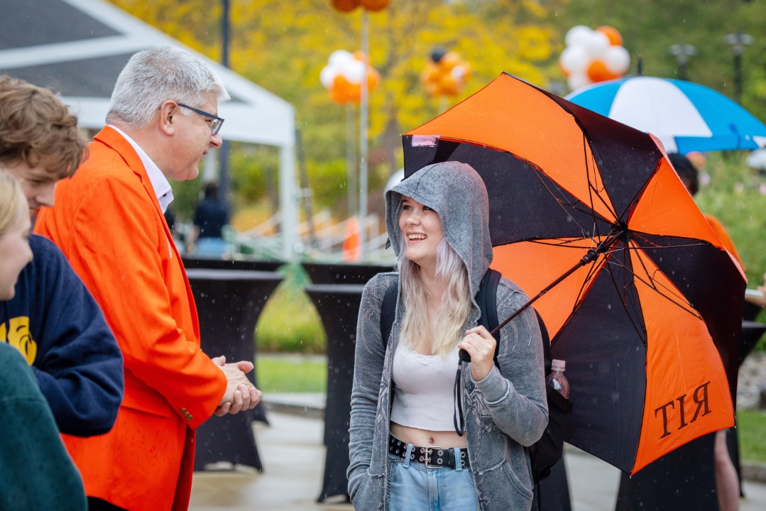 A student holding an orange and black RIT umbrella smiles while speaking with R I T president Sanders outdoors.
