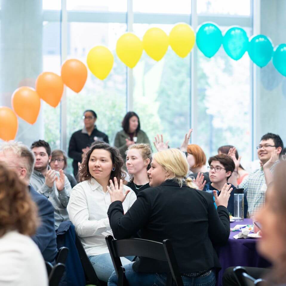 Several people sitting at tables with a rainbow made of balloons in the background.