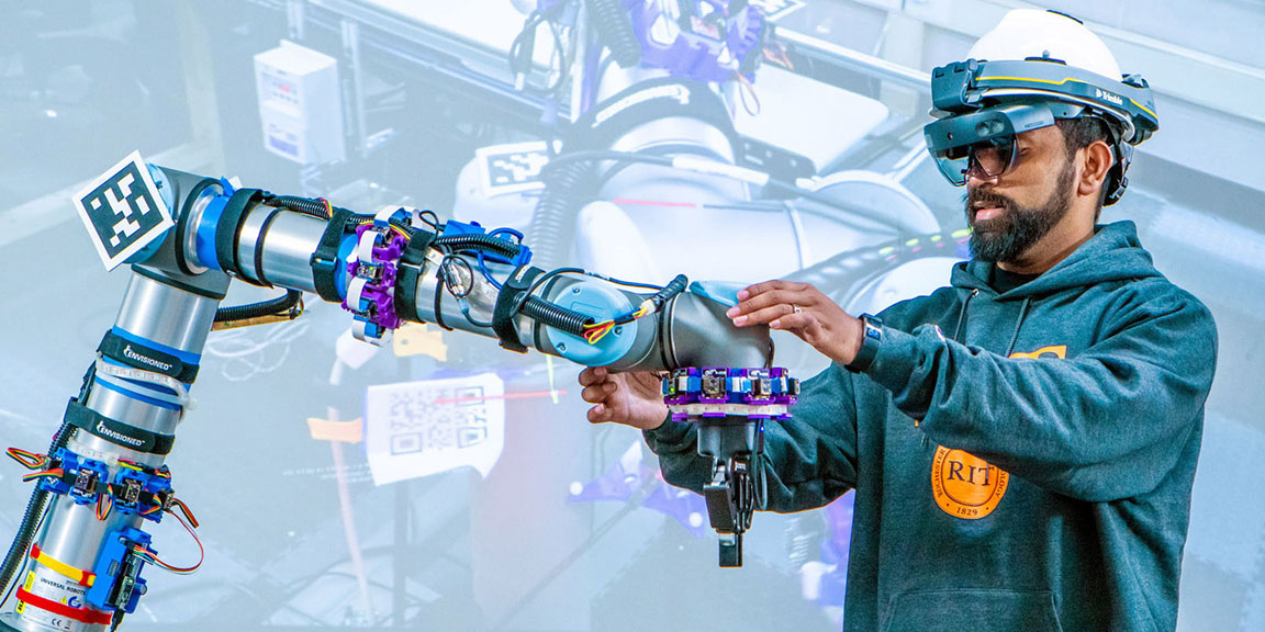 Man with a beard wearing protective hat/goggles working with a robotic arm.
