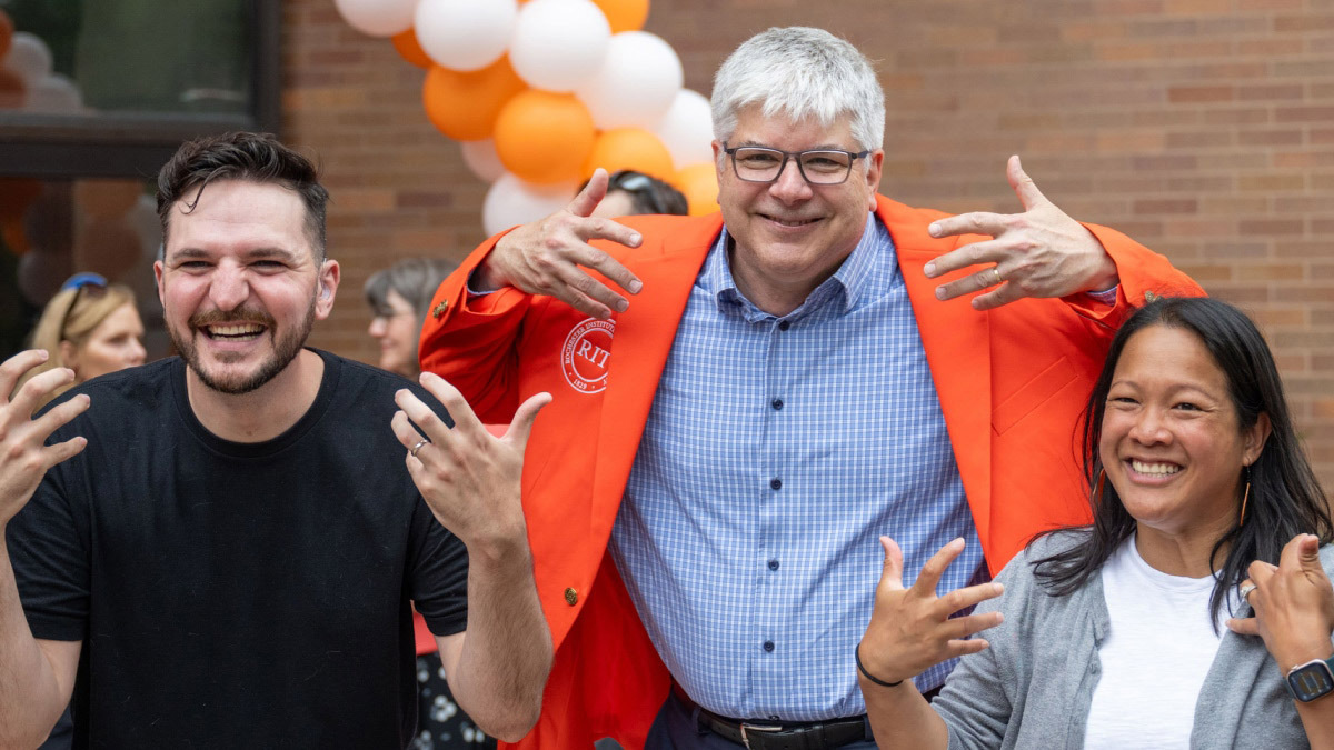 RIT President Sanders wearing an orange jacket, posing with 2 students.