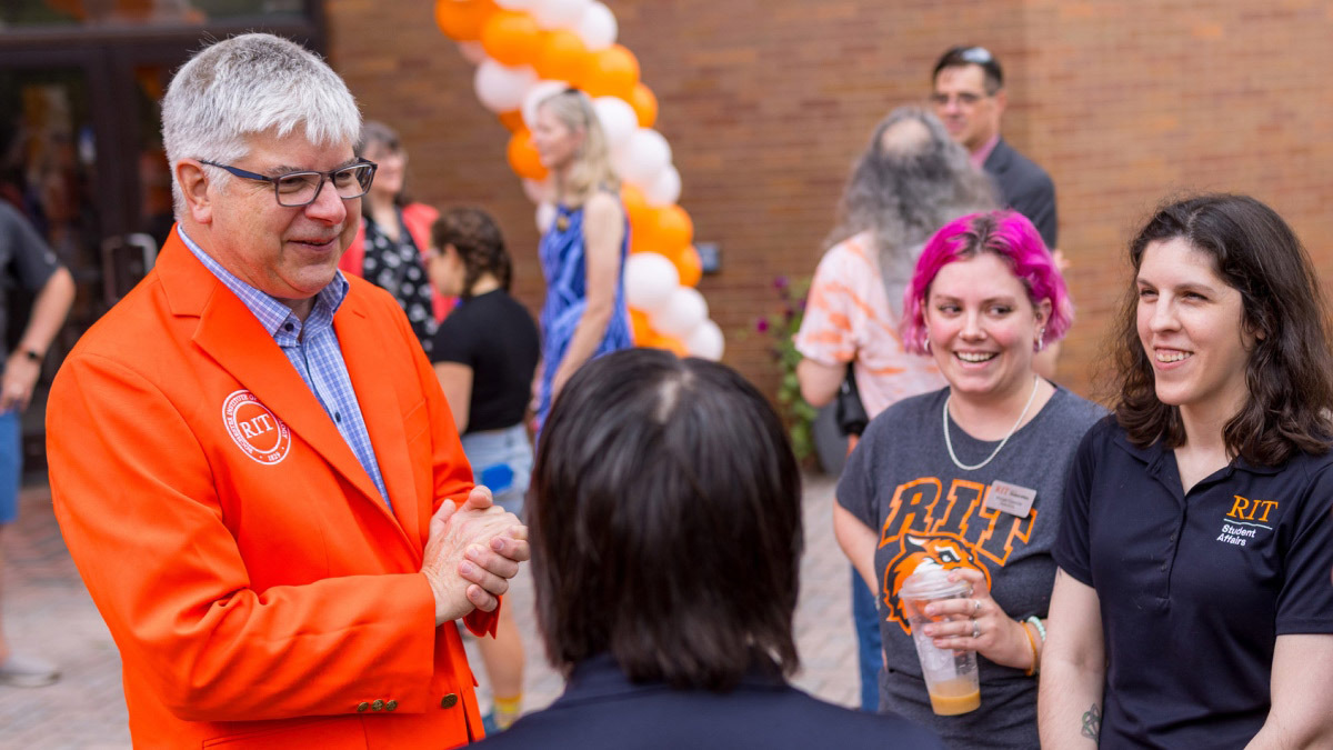 RIT President Sanders wearing an orange jacket, speaking with a group of people outside.