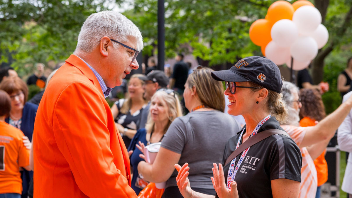 RIT President Sanders wearing an orange jacket, speaking to a person outside, near a cluster of orange and white baloons.