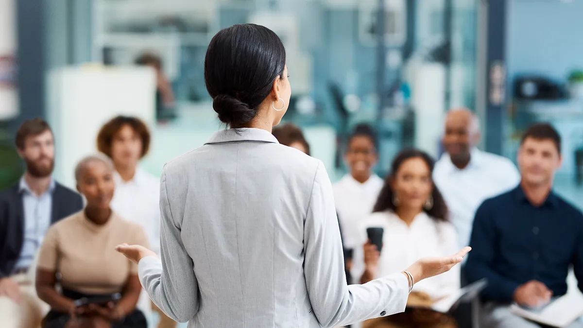A presenter stands facing an audience in a training or meeting room, gesturing while speaking. Several attendees sit and listen in the background.