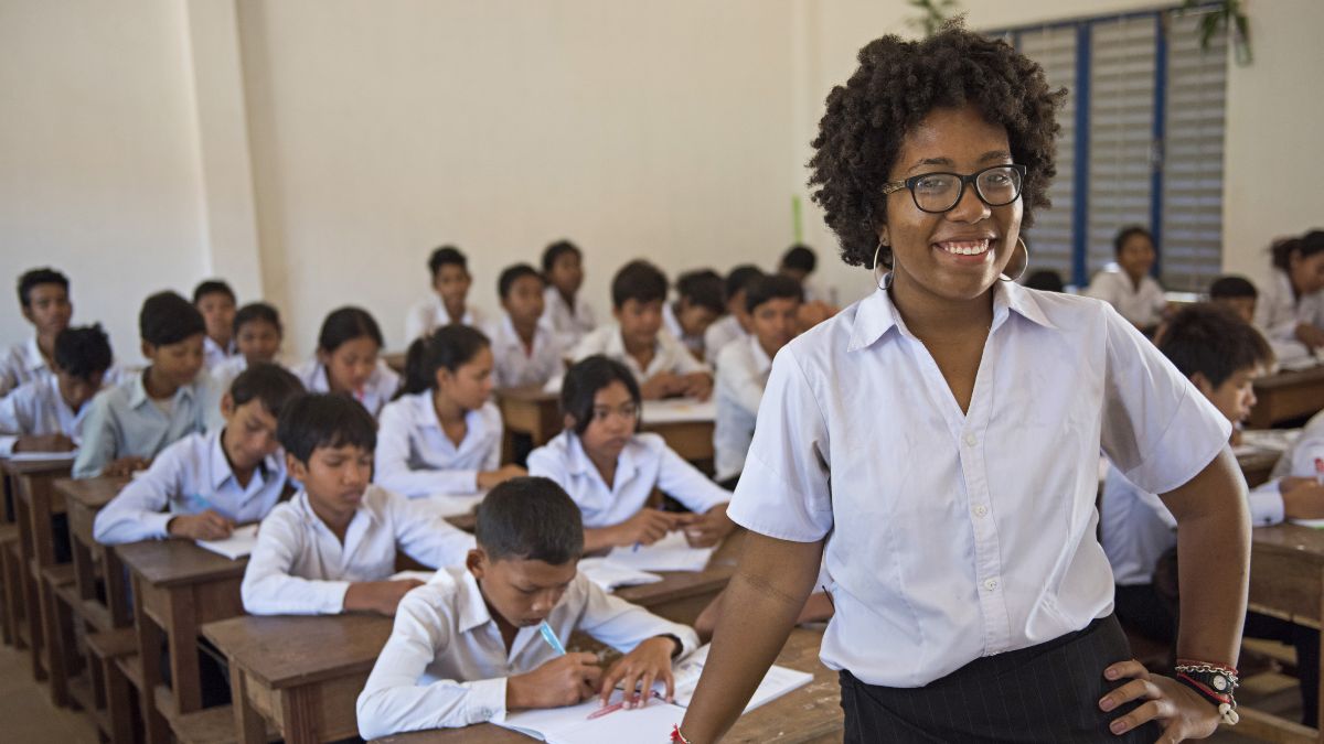 A teacher smiling in front of a classroom of attentive students in uniform.