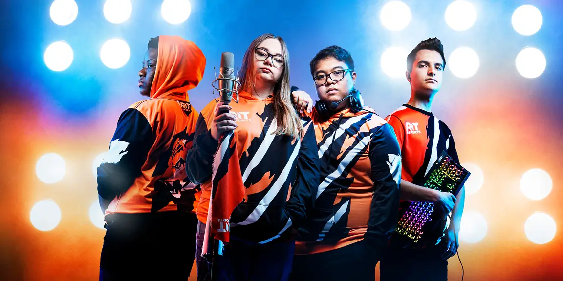 Four RIT eSports members, dressed in their uniforms, standing in front of stadium lighting, and holding gaming equipment.