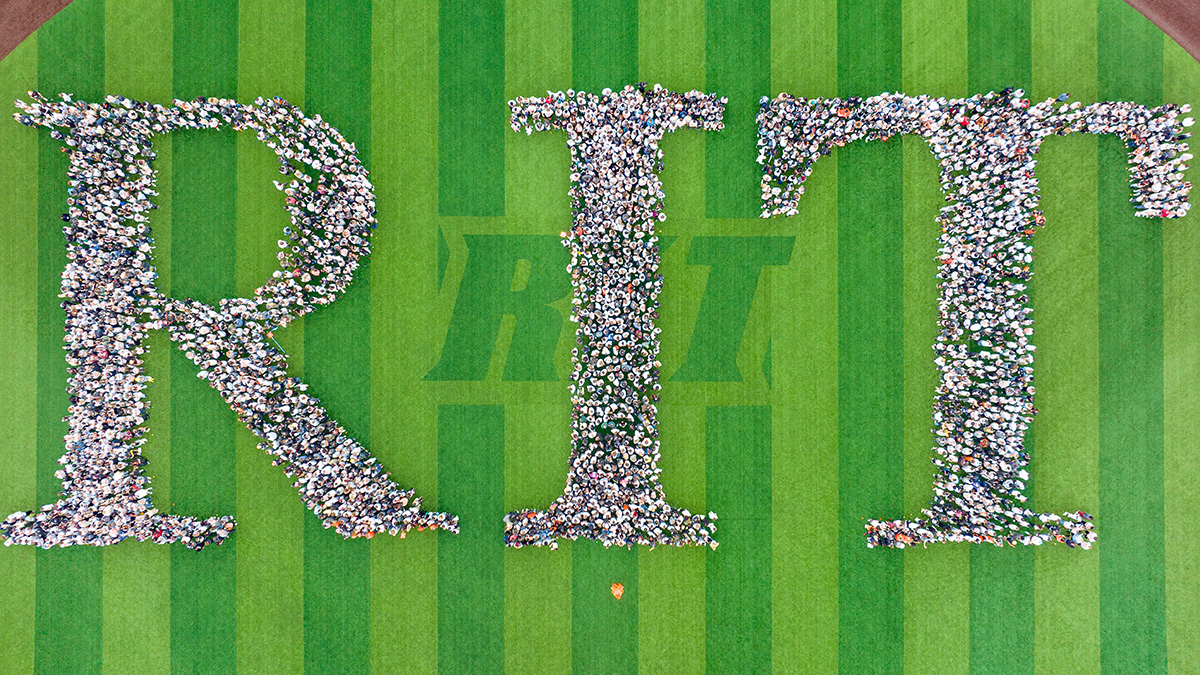 Letters R I T spelled out with people, taken from high above a grass field.