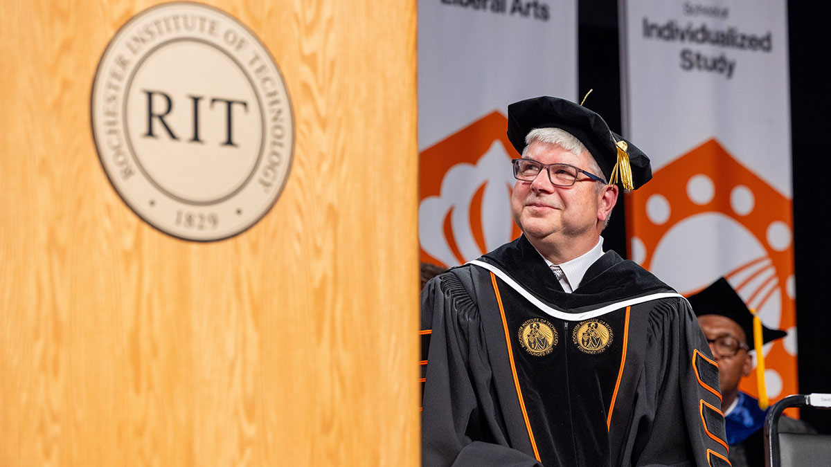 R I T president Bill Sanders wearing graduation regalia sitting on stage, with a podium with the R I T logo in the foreground.