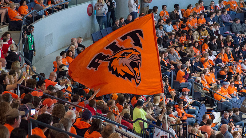An RIT student waving the RIT Tiger flag in the corner crew section at an RIT hockey game