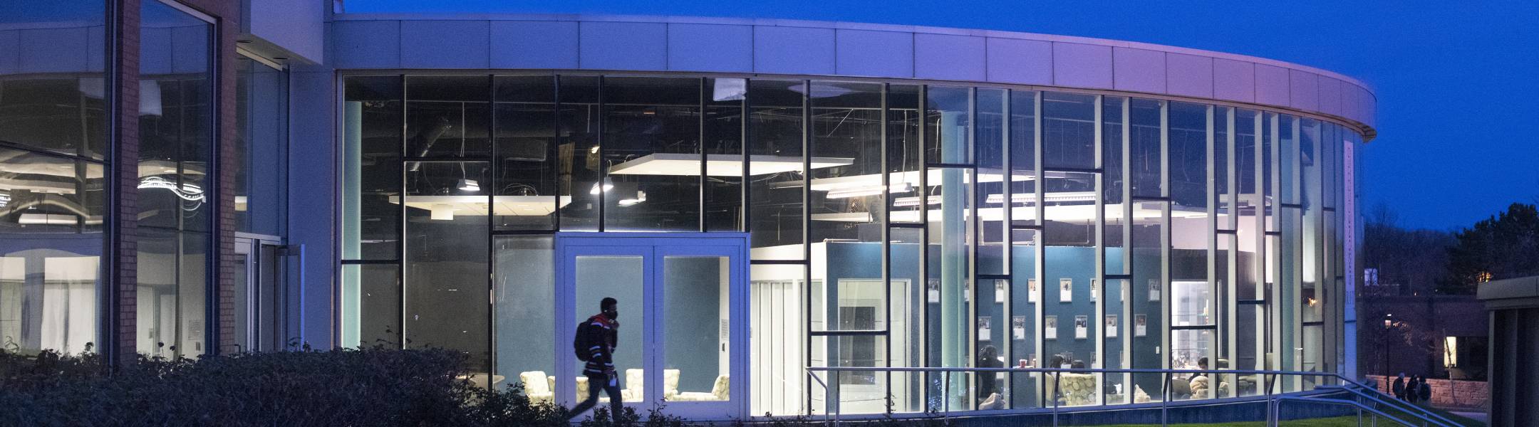 All glass building with lights on inside at night time with a student walking across the front