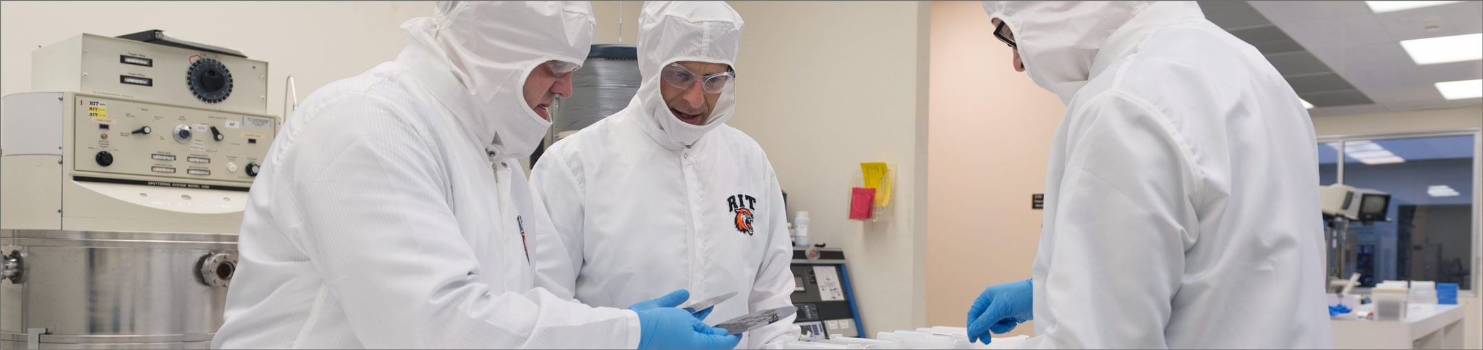 Three researchers in cleanroom suits looking at a disc of microelectronics.